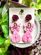 Marbled white and pink polymer clay earrings in the shape of a natural, curvy woman with a gold heart pendant, hanging from deep red rose posts. Earrings are showcased on a branded 'The Dappled Wood' card surrounded by lush greenery.