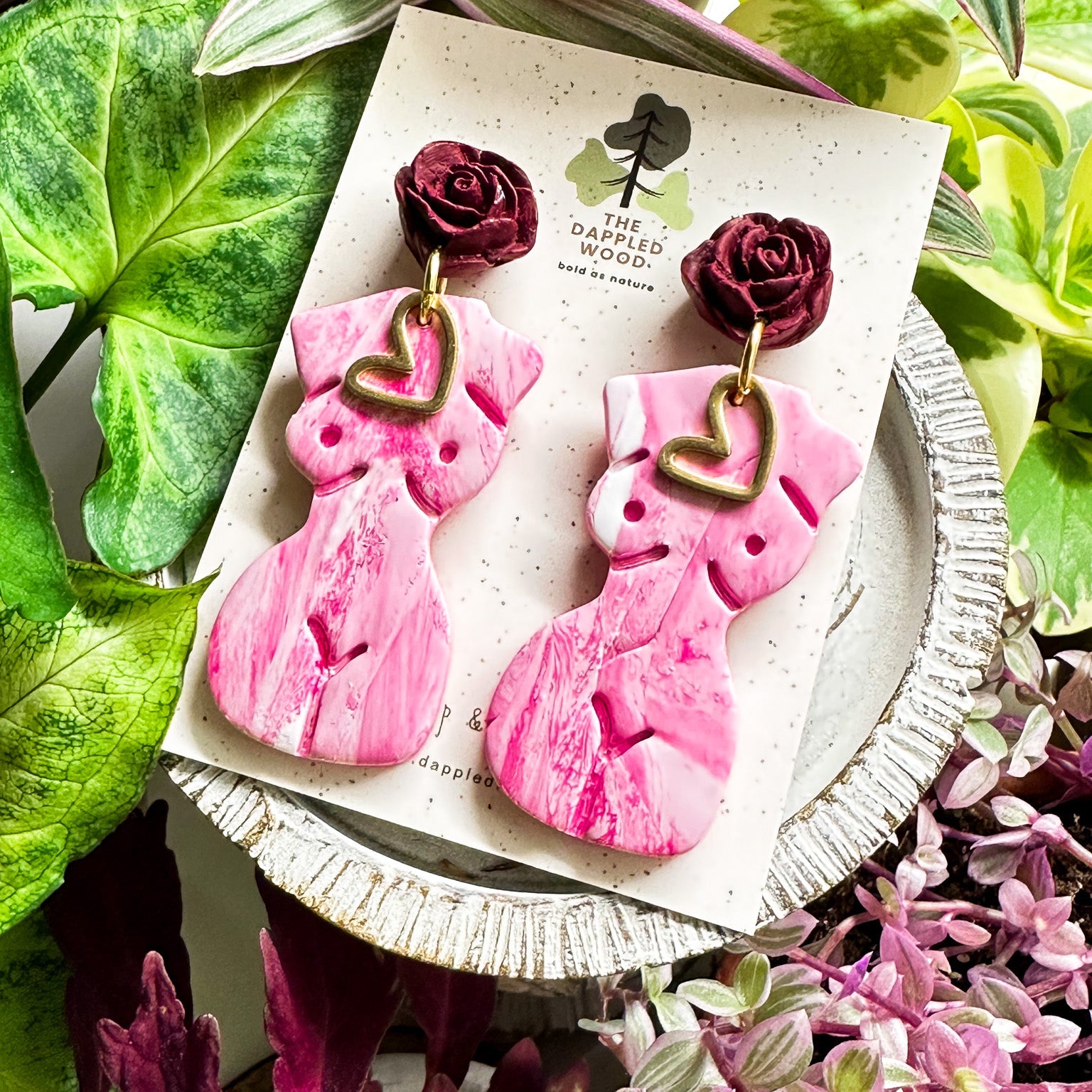 Marbled white and pink polymer clay earrings in the shape of a natural, curvy woman with a gold heart pendant, hanging from deep red rose posts. Earrings are showcased on a branded 'The Dappled Wood' card surrounded by lush greenery.