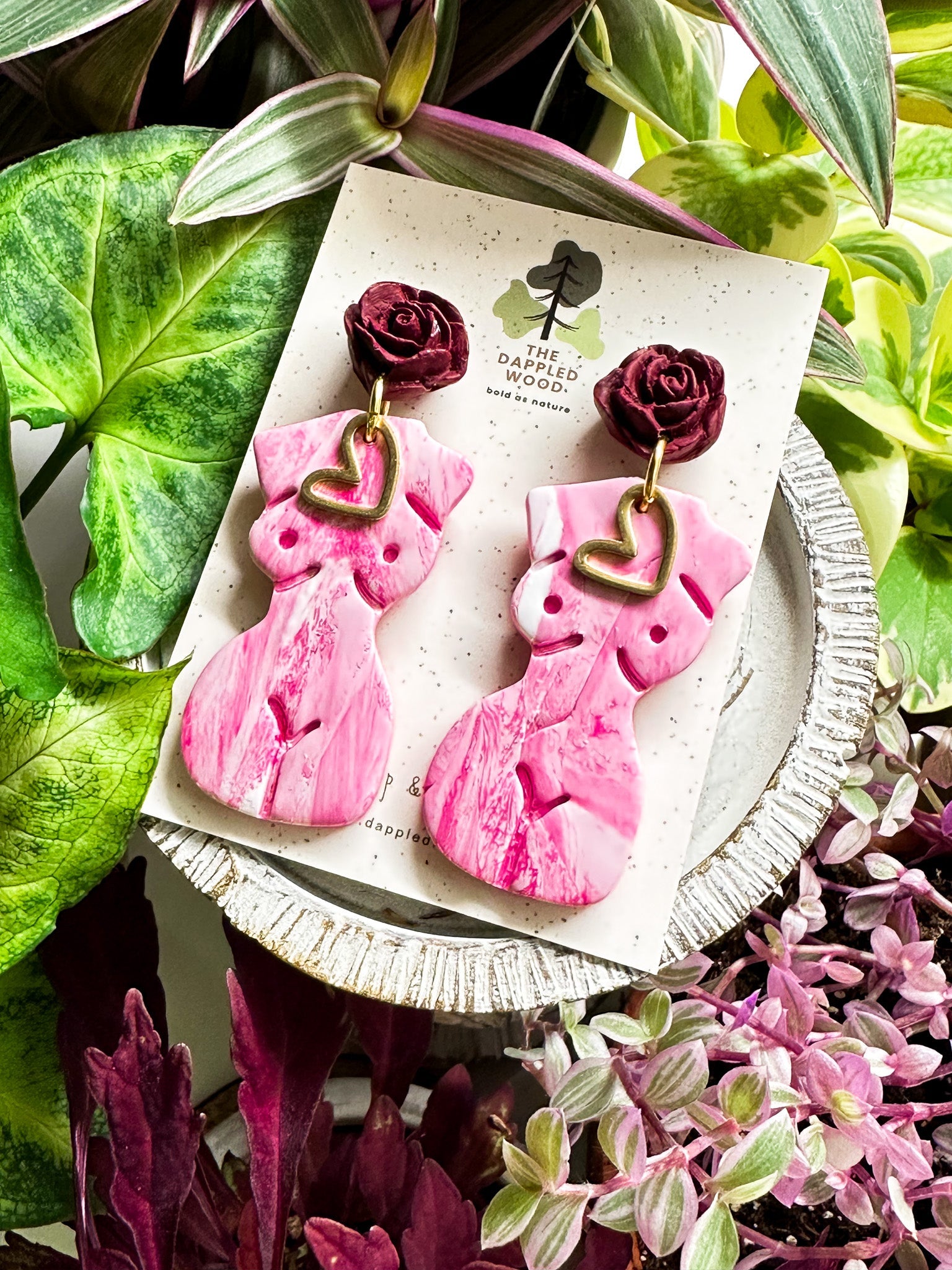 Marbled white and pink polymer clay earrings in the shape of a natural, curvy woman with a gold heart pendant, hanging from deep red rose posts. Earrings are showcased on a branded 'The Dappled Wood' card surrounded by lush greenery.