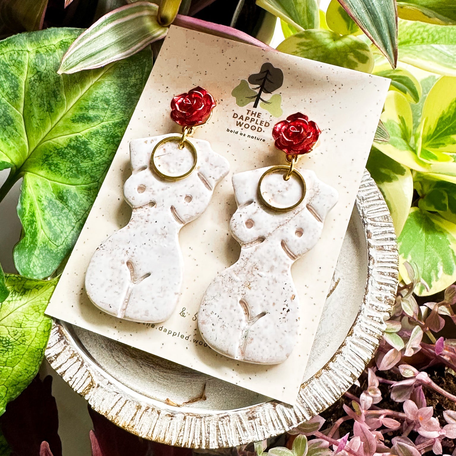 Elegant white polymer clay earrings with gold specks in the shape of a natural, curvy woman with a gold circle pendant, hanging from radiant red and gold rose posts. Earrings are showcased on a branded 'The Dappled Wood' card surrounded by lush greenery.