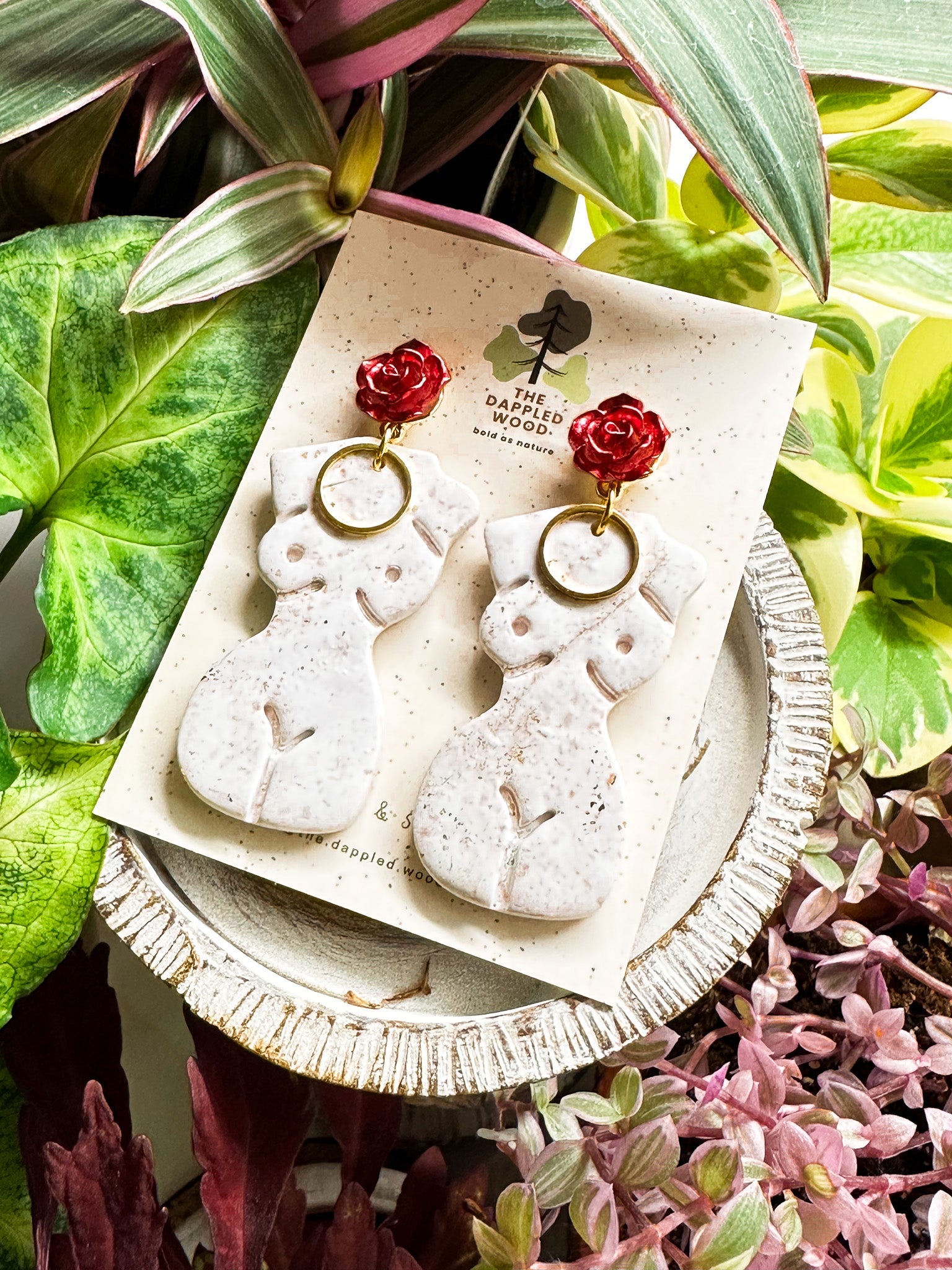 Elegant white polymer clay earrings with gold specks in the shape of a natural, curvy woman with a gold circle pendant, hanging from radiant red and gold rose posts. Earrings are showcased on a branded 'The Dappled Wood' card surrounded by lush greenery.