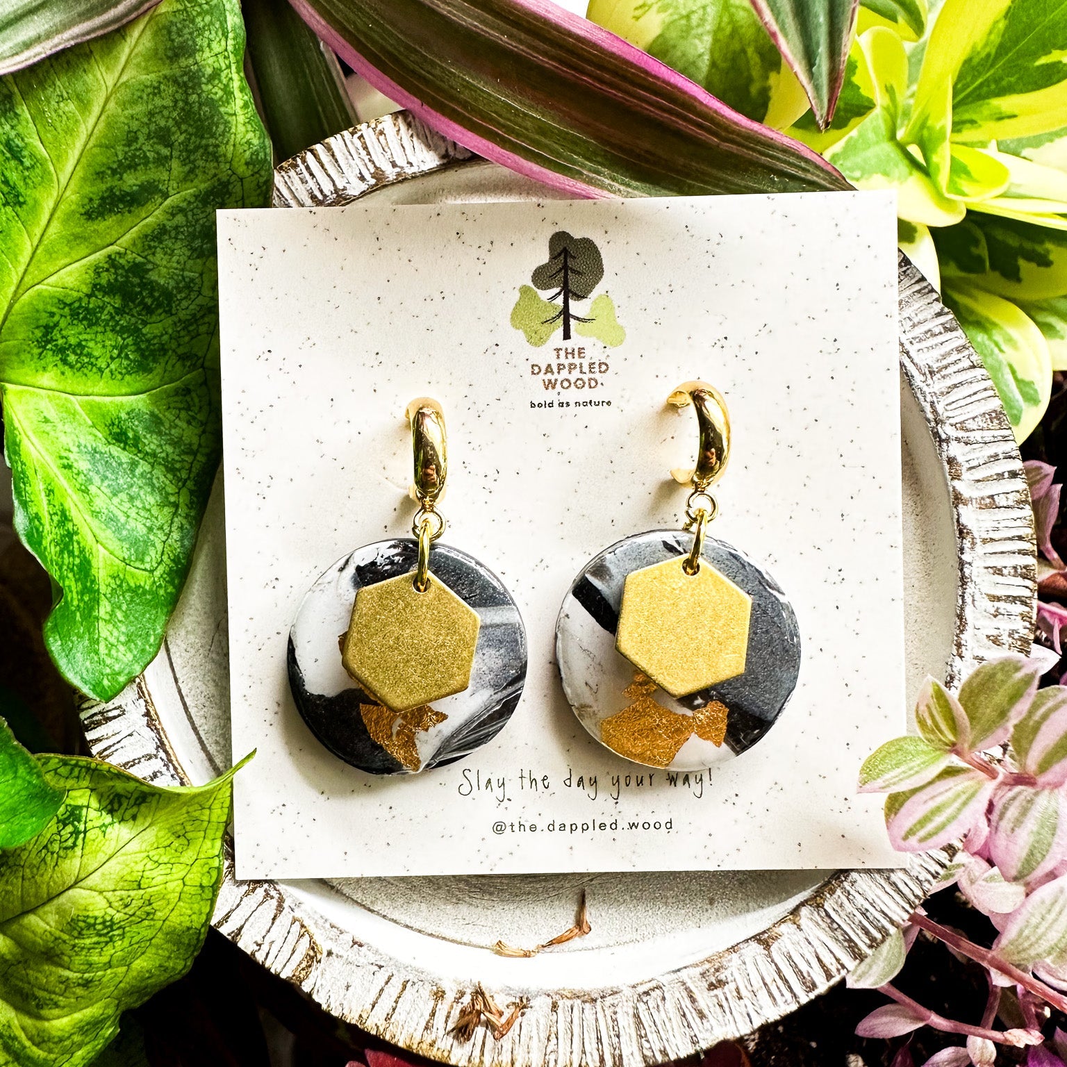 Gold, black and white  polymer clay earrings with brass hexagon charms dangled from gold hooks, presented on a speckled card with The Dappled Wood logo, amidst a lush background of green and pink plants.