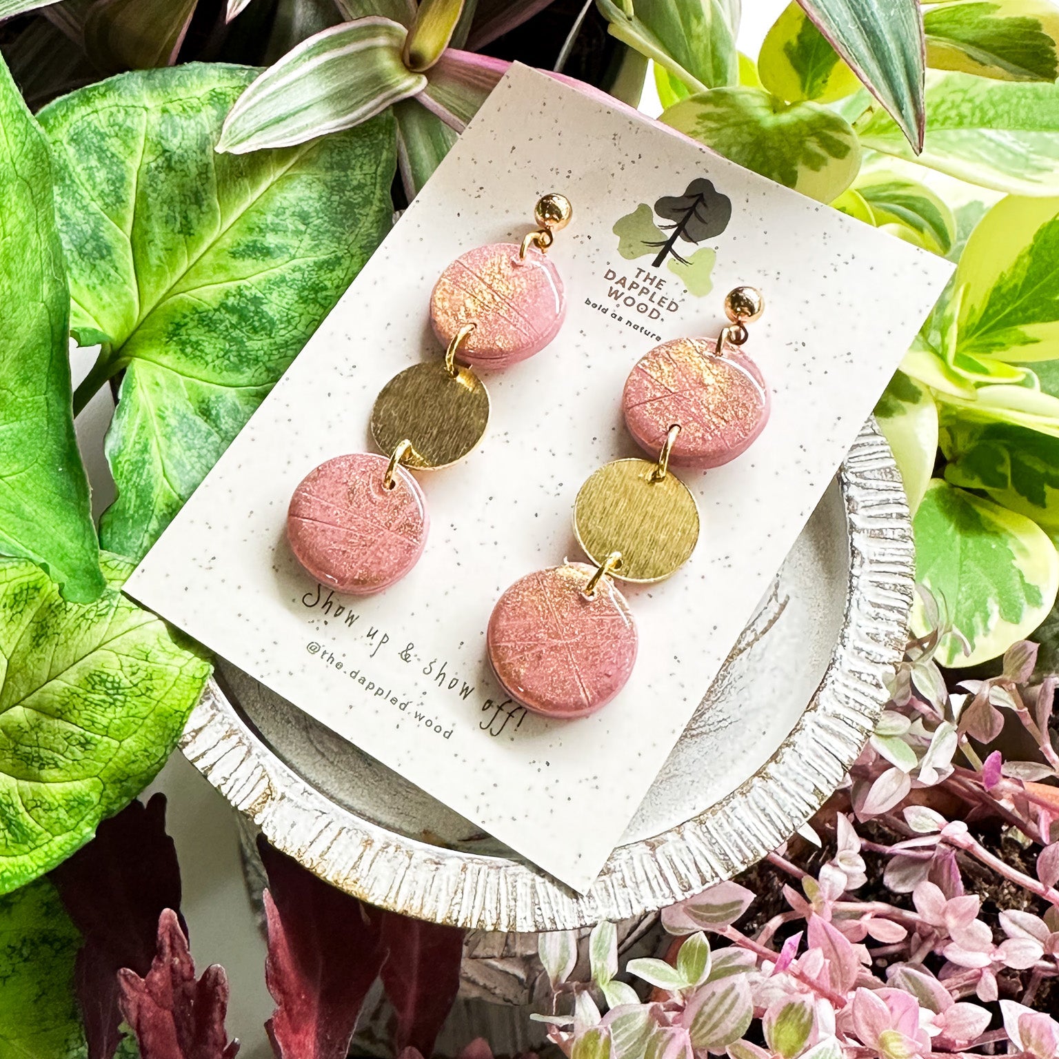 Handcrafted pink polymer clay disk earrings with gold shimmer and a textured brass connector charm, displayed on a speckled card with The Dappled Wood logo against a lush backdrop of green plants.