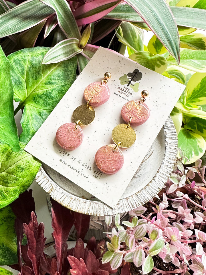 Handcrafted pink polymer clay disk earrings with gold shimmer and a textured brass connector charm, displayed on a speckled card with The Dappled Wood logo against a lush backdrop of green plants.