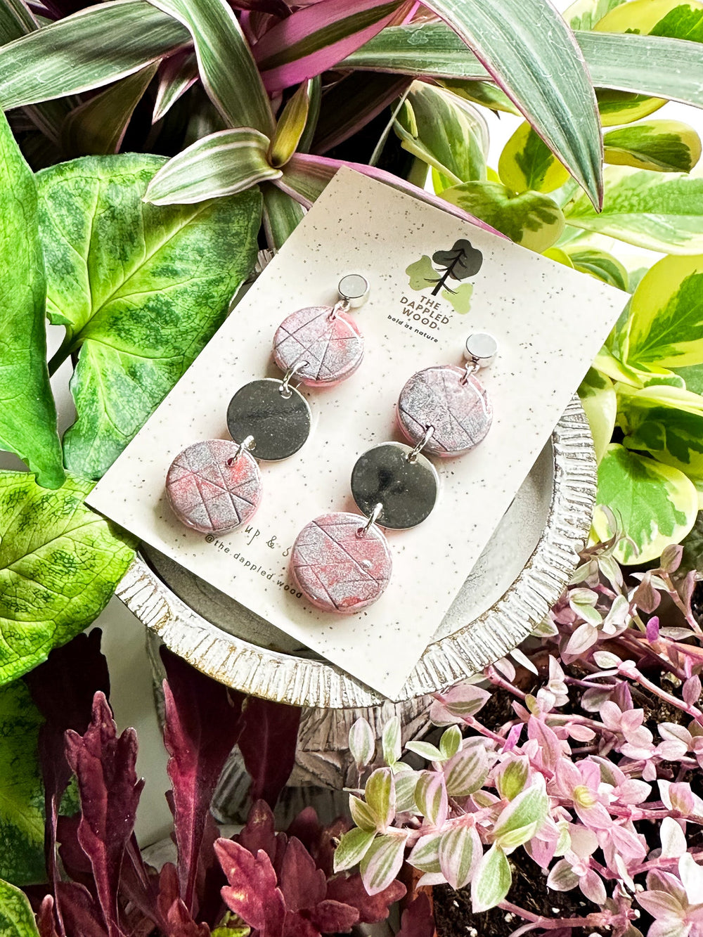 Handcrafted pink polymer clay disk earrings with silver shimmer and a smooth silver connector charm, displayed on a speckled card with The Dappled Wood logo against a lush backdrop of green plants.