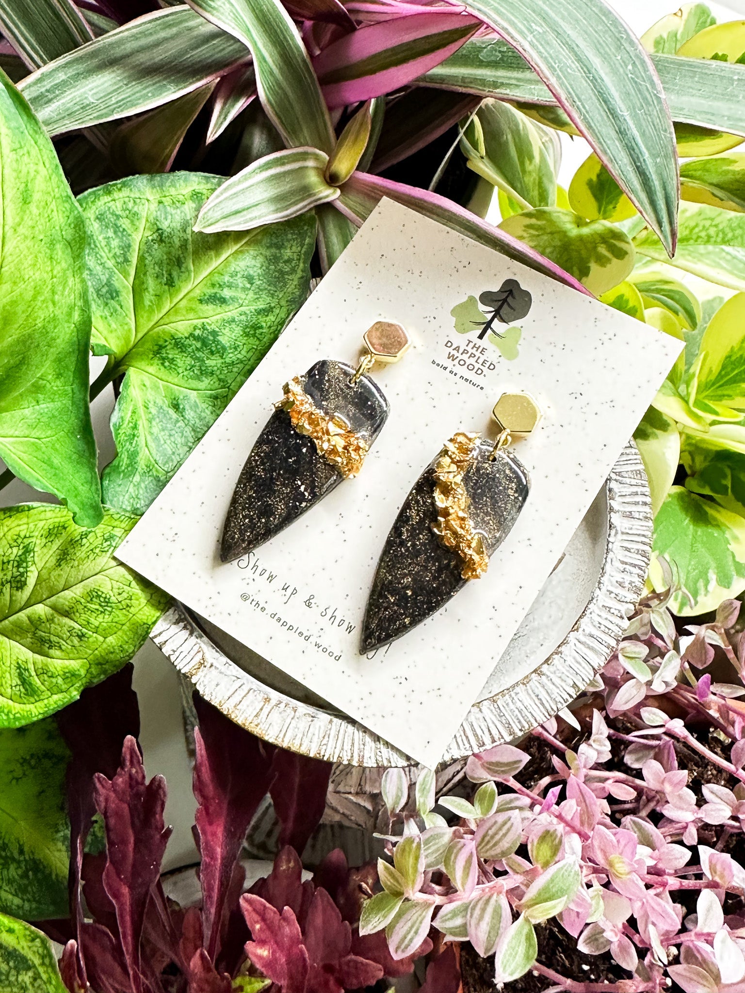 Black dagger shaped polymer clay 'Olivia' earrings with chunky gold glitter vein running through displayed on a speckled card with The Dappled Wood logo surrounded by green botanical background.