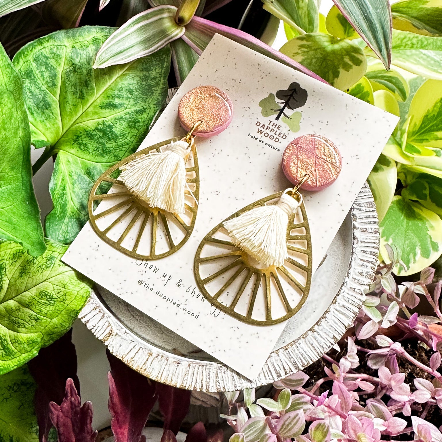 Handcrafted polymer clay earrings named 'Willa', showcasing a sparkling pink circular top with an openwork gold fan design and delicate tassels, presented against a speckled card with The Dappled Wood logo, surrounded by vibrant green and pink plants.